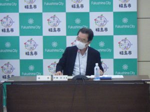 長テーブルの前に着席した経営審議会会長が福島市のバックボードの前で話をしている様子の写真