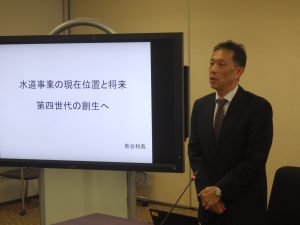 スーツ姿の男性が、会議室で左奥に「水道事業の現在位置と将来第四世代の創生へ」とディスプレイに表示されたスライドを背景に立って話をしている様子の写真
