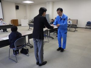 室内で、スーツ姿の男性が青い作業服を着た男性に委嘱状を渡している様子の写真