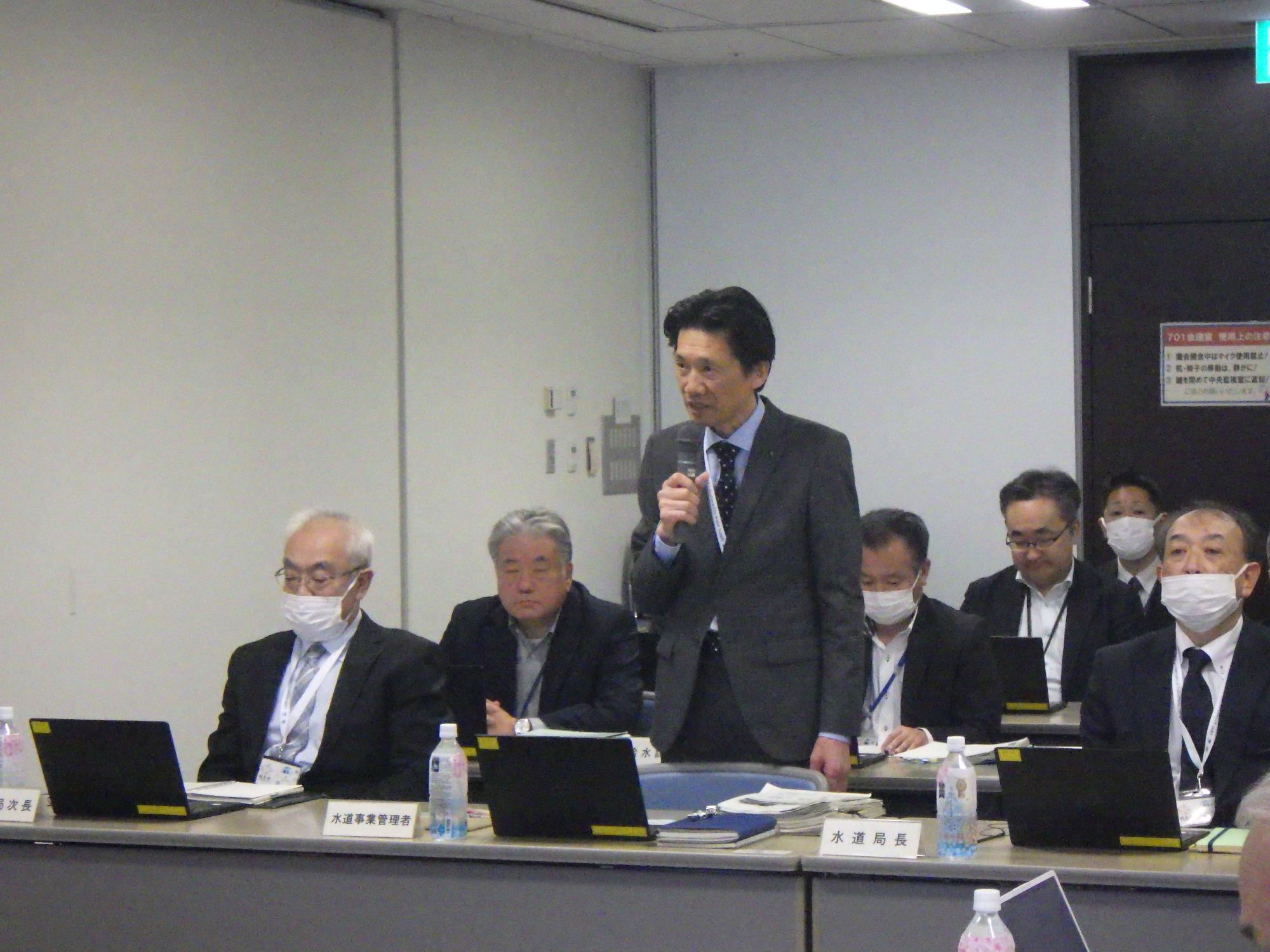 室内で、スーツ姿の関係者の男性が中央に立ってマイクを持ち発言しており、周囲のテーブル席では複数の関係者がノートパソコンや資料を前に着席している様子の写真