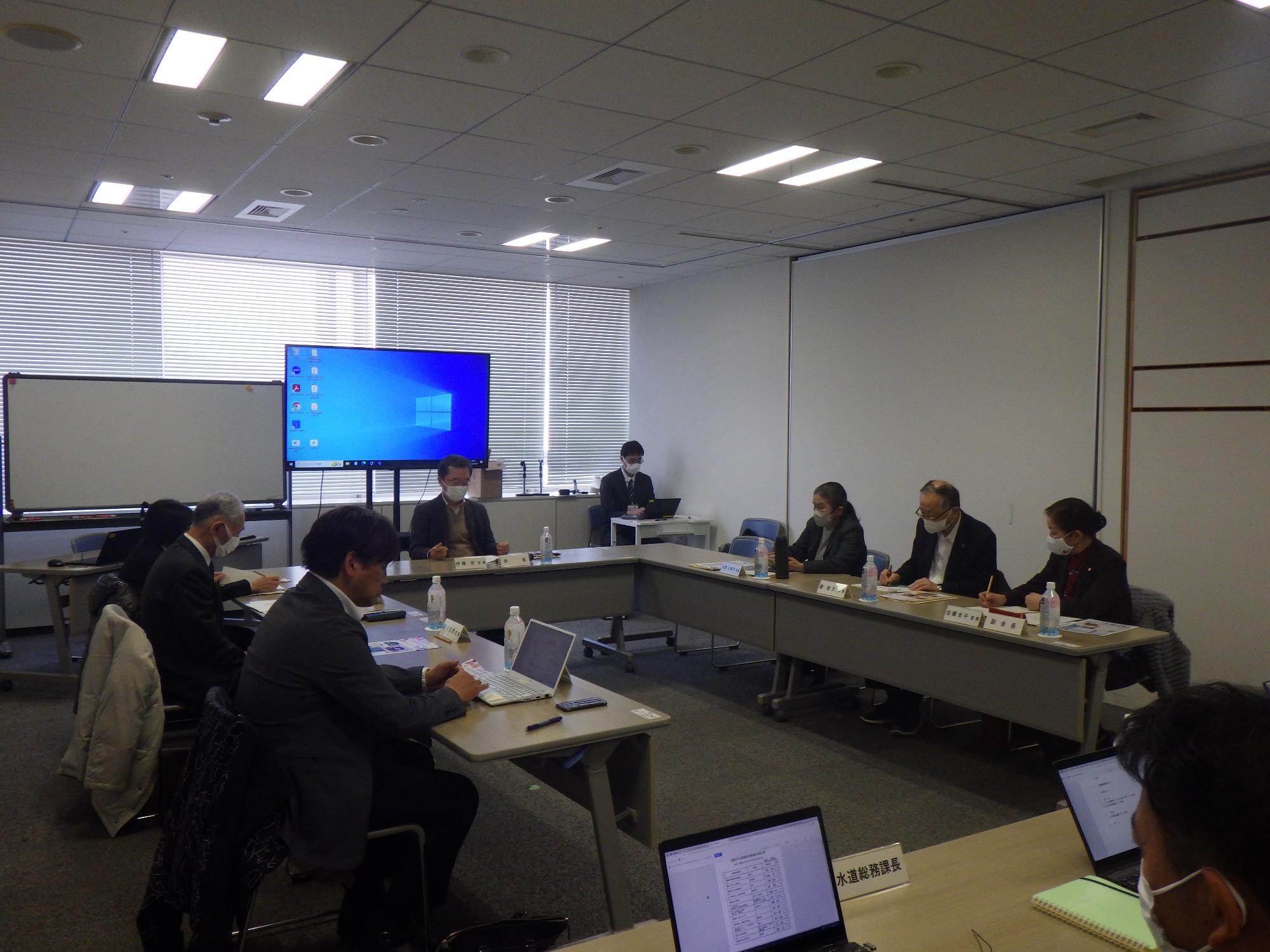 広い会議室で、長テーブルをコの字型に配置し、十数名の人々が着席した会議の様子の写真