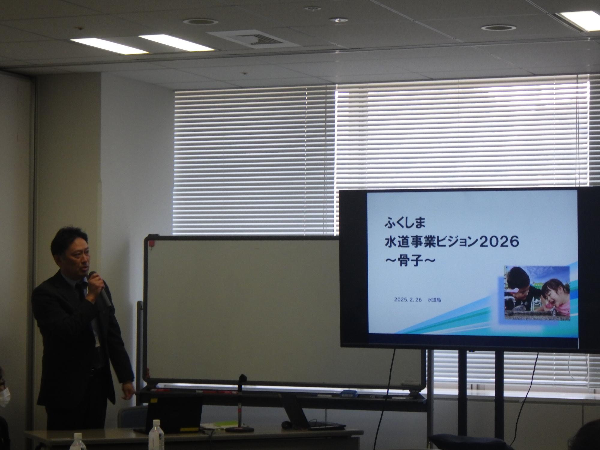 会議室で、スーツ姿の関係者の男性がマイクを持って立ち、その右側にあるディスプレイには「ふくしま水道事業ビジョン2026～骨子～」というタイトルのスライドが表示されている写真