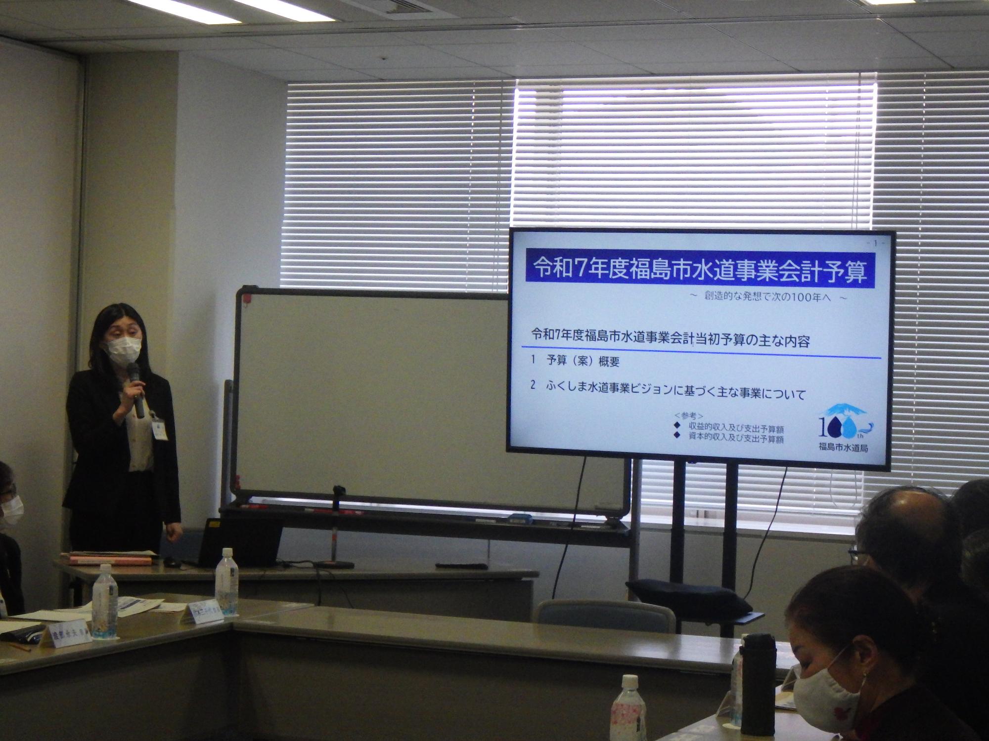 会議室で、スーツ姿の関係者の女性がマイクを持って立ち、右側のスクリーンには「令和7年度福島市水道事業会計予算」というタイトルと主な内容が表示されているプレゼンテーションの様子の写真
