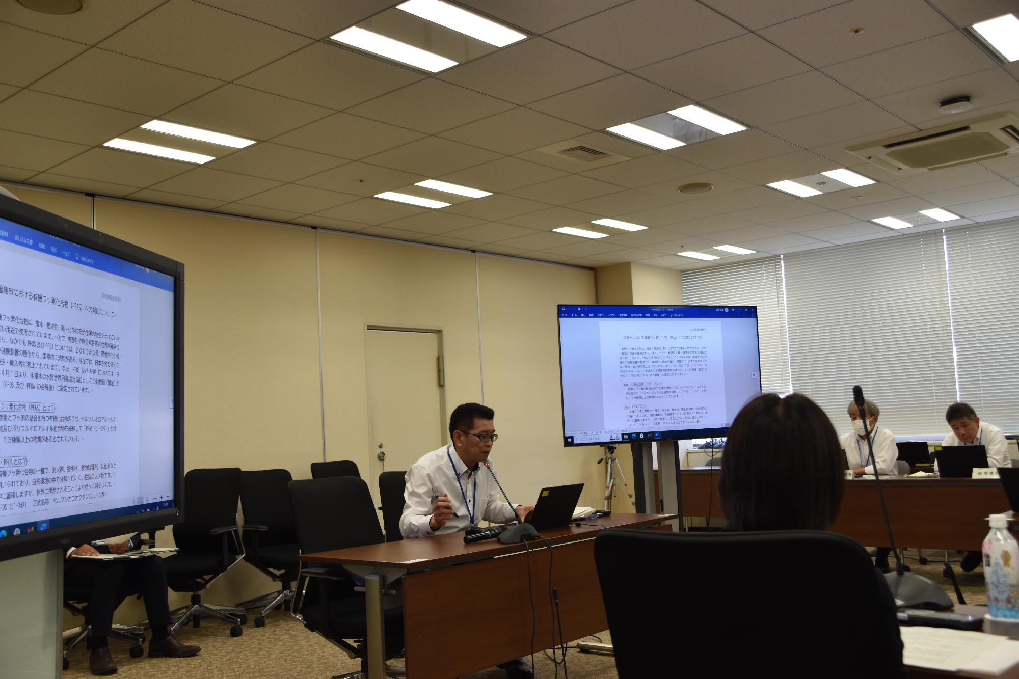 広々とした会議室で、中央の長机に座った白いシャツの男性が、前方のモニターに投影された資料を参照しながら発言している様子の写真