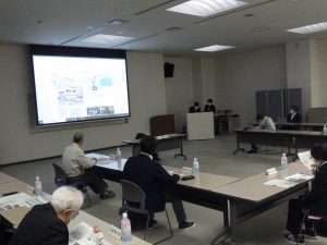 U字型に配置されたテーブルに数名の参加者が座っており、スクリーンには地図や写真などの画像を含むプレゼンテーション資料が投影されている会議風景の写真