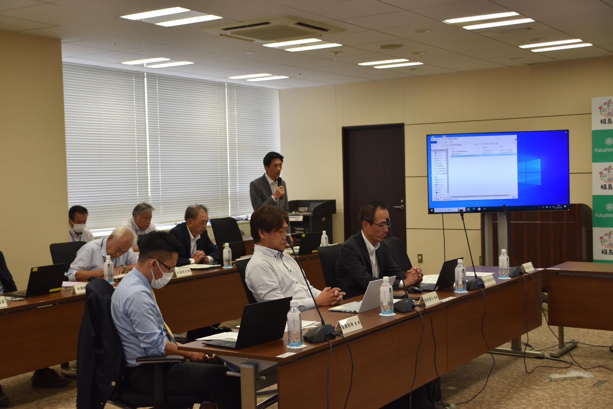 長テーブルに関係者が複数名座り、部屋の奥の中央に立ったスーツ姿の人物がマイクを使って発言している様子の写真