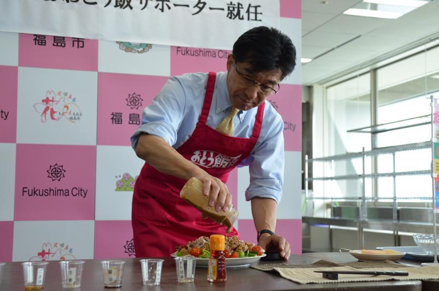 赤いエプロンをした市長がドレッシングのような物を料理にかけている様子を写した写真