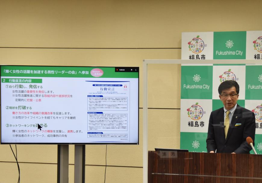 福島市のバックボードの前に市長が立ち、モニターに資料を写しながら定例記者会見を行っている様子の写真