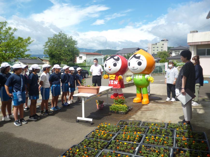 小学生が整列してその前に机が置かれプランターが置かれその横で男性と人KEN(じんけん)まもる君、人KEN(じんけん)あゆみちゃんが並んで贈呈式が行われている写真