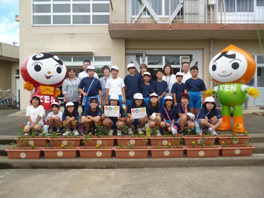 階段にプランターを並べ、その後ろで子供たちが人KEN(じんけん)まもる君、人KEN(じんけん)あゆみちゃんと一緒に記念撮影をしている写真