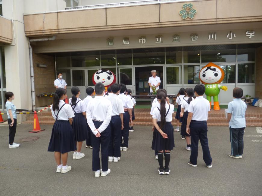 校舎前で1人の男性が説明をし、子供たちが整列して話を聞いている後方から写した写真