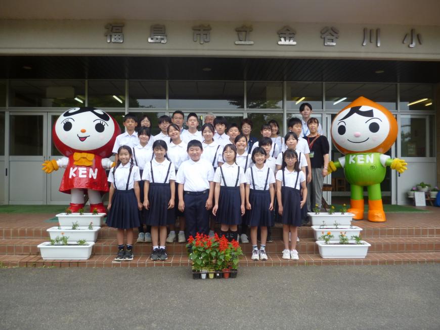 校舎前の階段に4列に並んだ子供たちと両端に並んでいる人KENまもる君と人KENあゆみちゃんとの記念撮影をしている写真