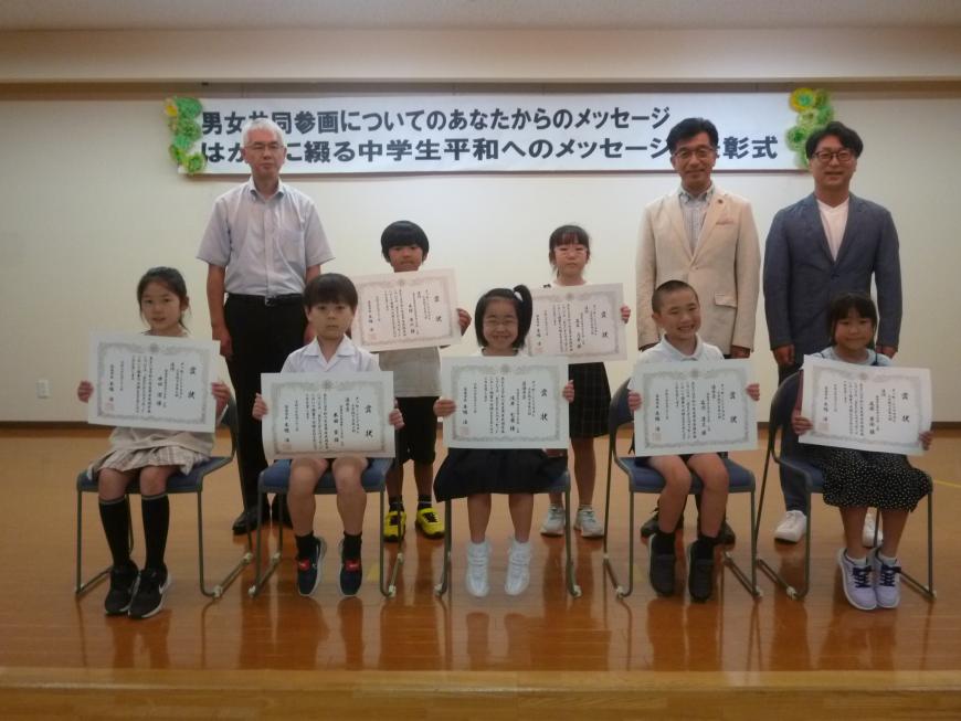 関係者3名と1列目の椅子に5名の小学生、中央に2名の小学生が賞状を見せながら笑顔で記念撮影をしている写真