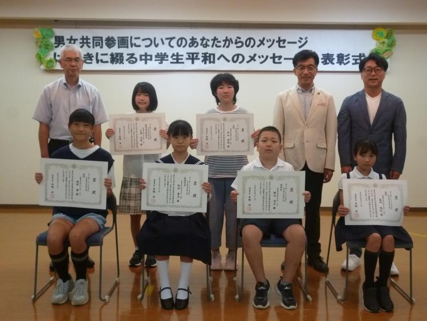 関係者3名と1列目の椅子に4名の小学生、中央に2名の小学生が賞状を見せながら笑顔で記念撮影をしている写真
