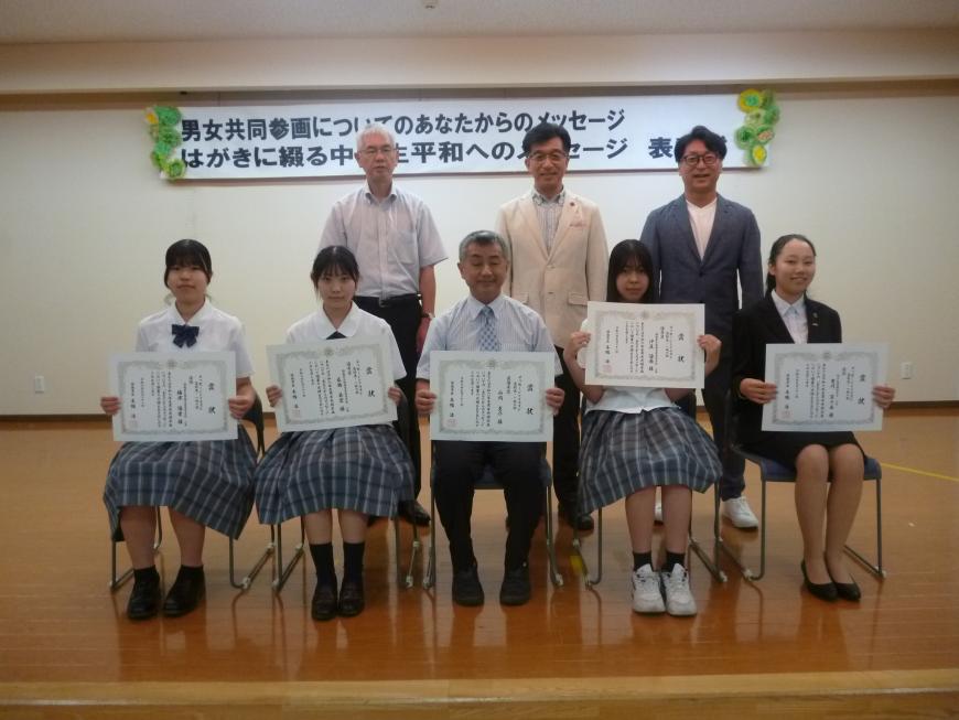 関係者3名が2列目に並び、中央に男性1名、その隣に2名ずつ女子高生が座り賞状を見せながら笑顔で記念撮影をしている写真