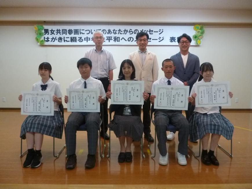 関係者3名が2列目に並び、中央に女性1名、その隣に各1名ずつ男子学生、その横に1名ずつ女子学生が座り賞状を見せながら笑顔で記念撮影をしている写真