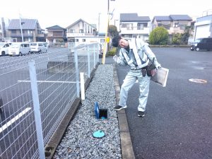 舗装された駐車場の隅で、作業服と反射ベストを着用した男性が、砕石が敷かれた側溝内の青い小さな蓋が開いた水道メーターを覗き込んでいる写真