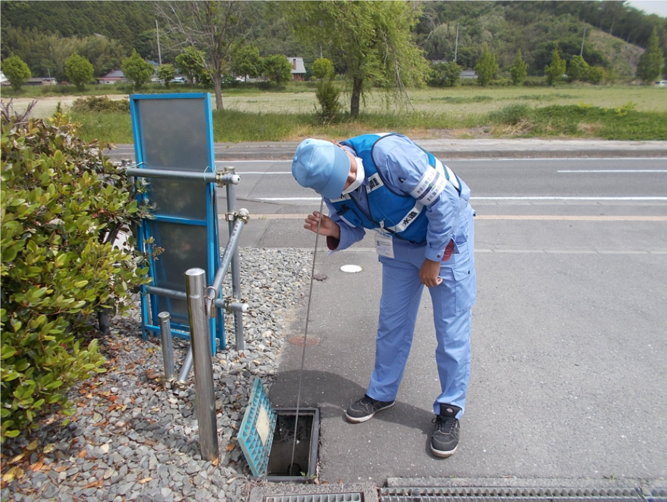作業服と反射ベストを着用した男性が、水道メーターの青い蓋を開けて調査している写真