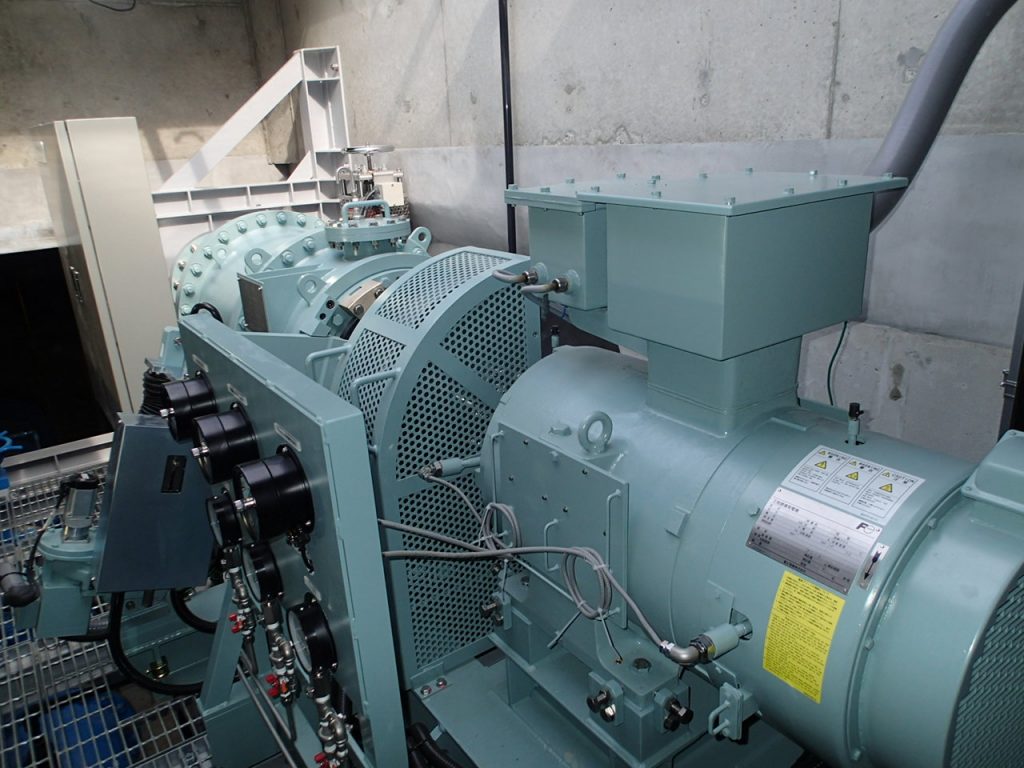 コンクリートの壁に囲まれた流入弁室内で、配管や電気配線が接続された金属製の装置が並ぶ水車発電機を写した写真
