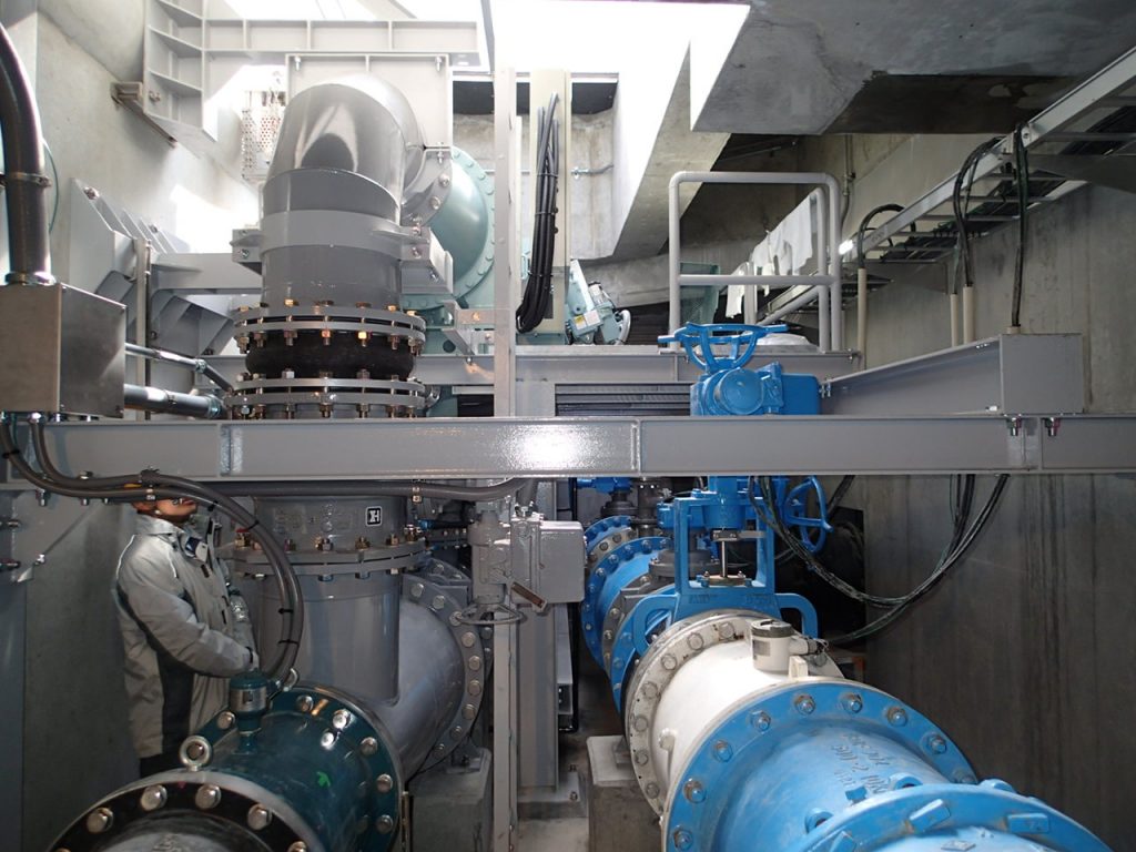 作業服を着た作業員が点検を行う中、灰色と青の大口径の配管が複雑に配置され、フランジで接続された管路や青いバルブが並ぶ流入弁室内の配管を写した写真