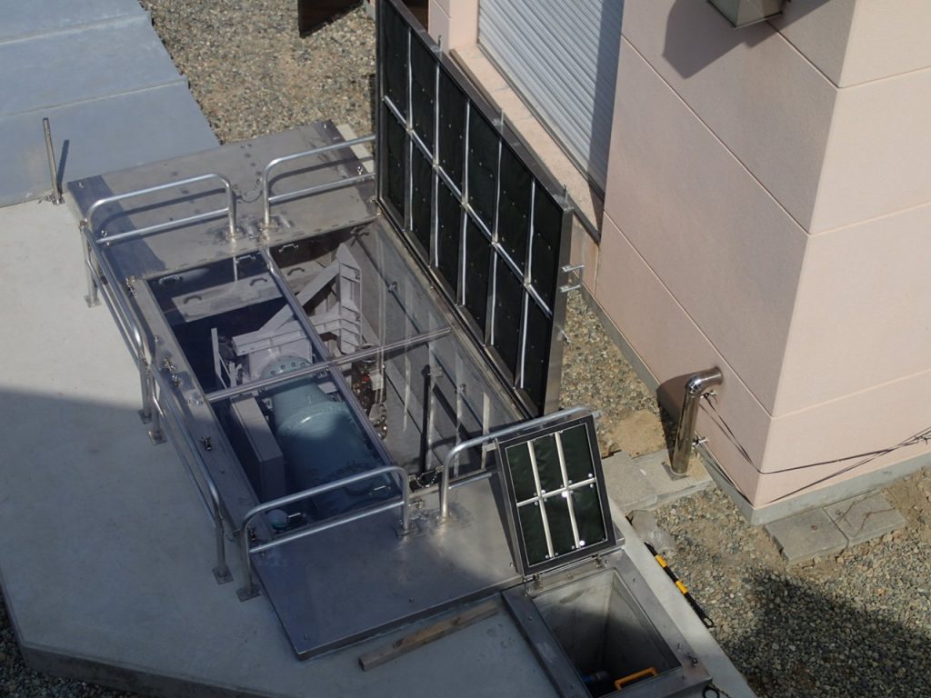 金属製のハッチが開けられており、その内部には青い大きな機器や配管の一部が見え、周囲は手すりで囲まれ安全対策が施されている様子を北部配水池屋上から撮影された流入弁室のマシンハッチの写真