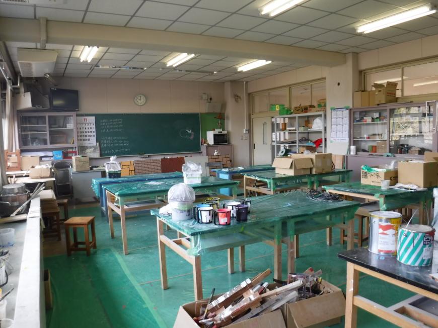 前方に黒板があり作業する台が6つ置かれペンキの缶などが置かれた塗装科実習室の写真