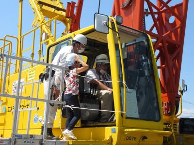 工事現場で使用する黄色い自動車に作業員の方が座っており、見学の女の子が車の横に立ち説明を聞いている様子の写真