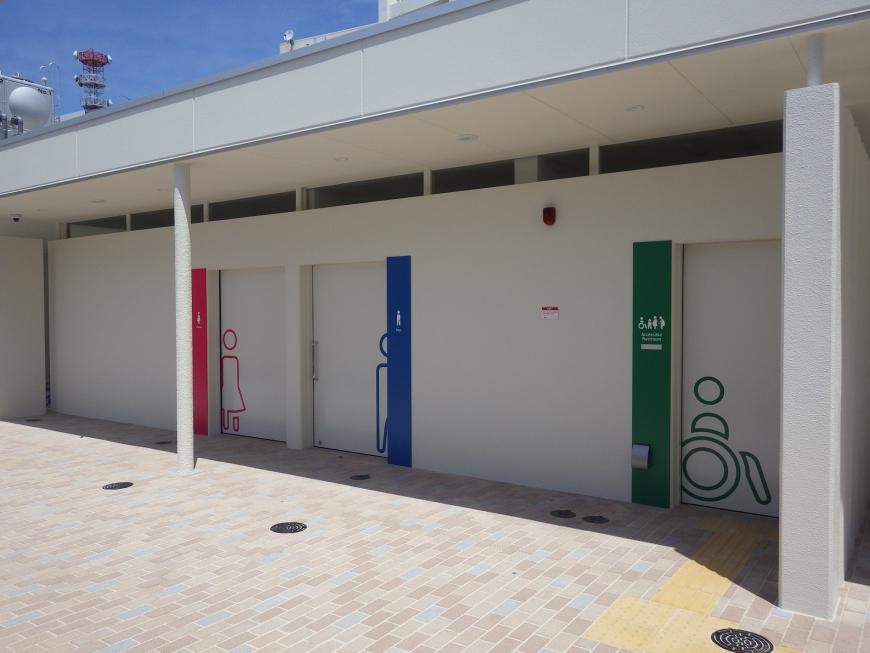 白を基調とした外壁で左からピンク色の女性用トイレと青色の男性用トイレ、緑色のバリアフリートイレのドアを撮影した写真