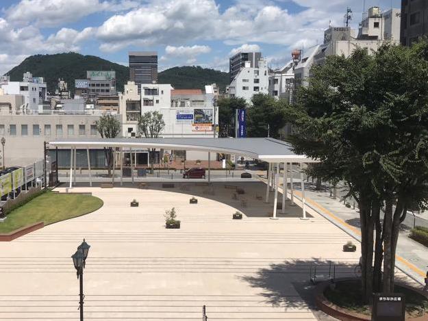 円形の花壇に木が植えられたビル群に囲まれた広場の奥にあるL字型の大きな屋根が設置された東屋を高台から撮影した写真