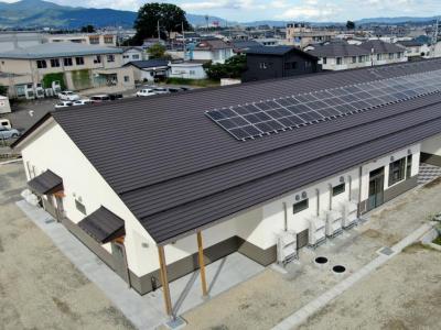 白壁で太陽光パネルが設置された横長の黒色の屋根を、周囲の山や住宅が見渡せる高台から撮影した写真