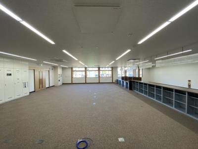 左側に出入り口の扉、奥に太陽光が差し込む窓ガラス、右の壁側に棚が設置された縦長の室内の写真
