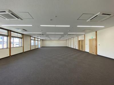 左側に太陽光が差し込む窓ガラス、右側に出入り口の扉が設置され、グレー色絨毯が敷かれた縦長の室内の写真