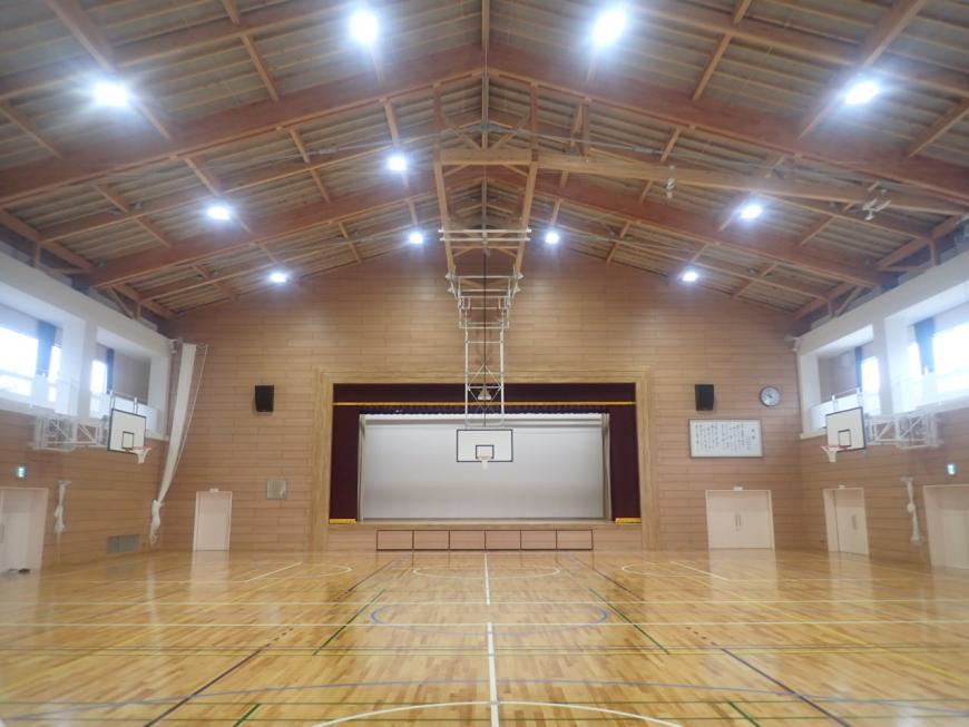 左右の壁側にスピーカーが設置されたステージがあり、天井に設置された照明の光が床に反射した体育館内の写真