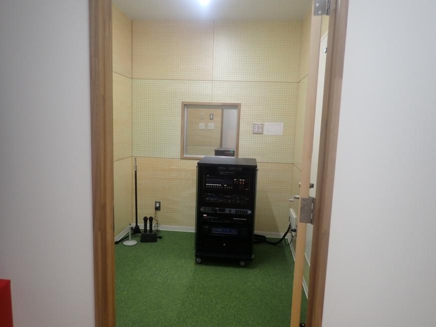 奥に隣の部屋が見える正方形の窓ガラスが設置され、2つのマイクと音響機器が設置された室内の写真