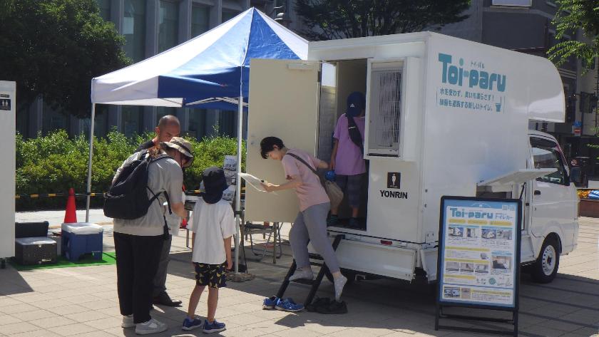 赤い車体の救助工作車が展示されており、親子連れが見学に訪れ、係の男性の側で男児が重そうな機械を両手で持っている様子の写真