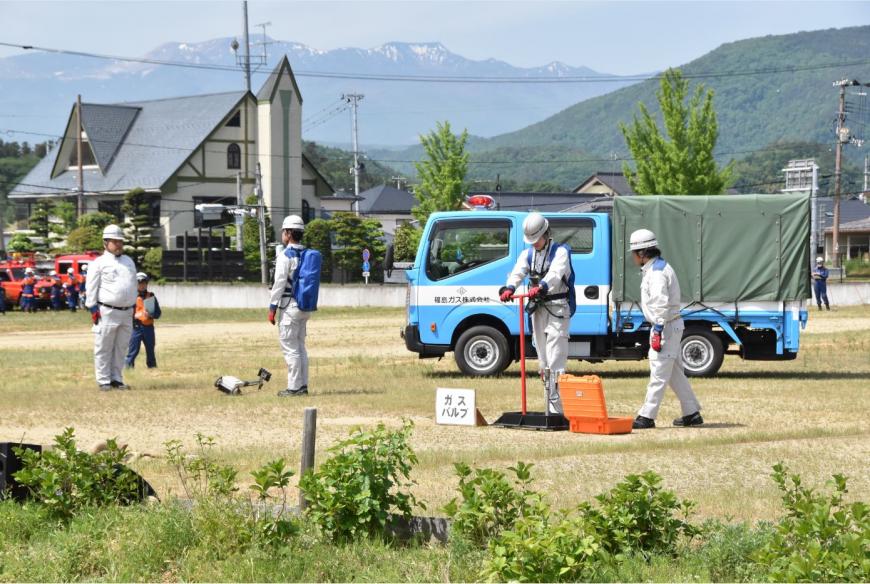 「ガスバルブ」と書かれた紙が置かれた横でヘルメットを被った男性がT字の形をした道具を使い作業を行っている様子の写真