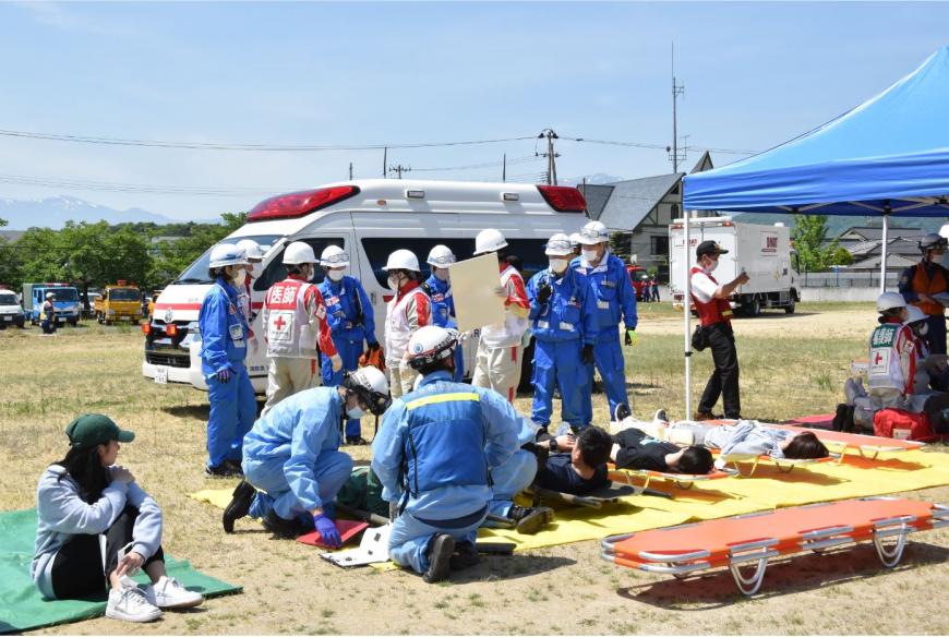 救急車がとまっている横に敷かれた黄色いシートの上に4名ほどの方が担架に乗せられ横になっている写真