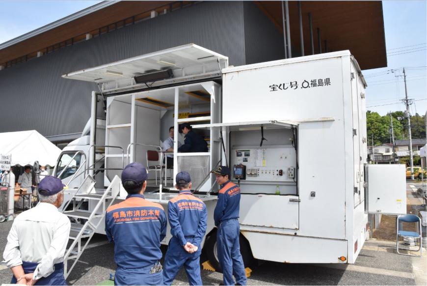 全体が白いトラック荷台部分の後方に機械のパネルのような物が設置され、荷台の前方部分には階段が設置され男性が座るスペースがあるトラックの写真