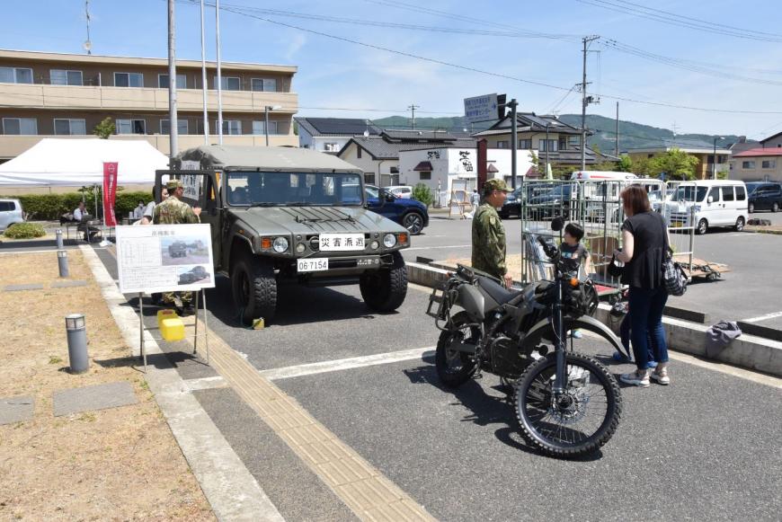 前方に「災害派遣」と書かれた自衛隊車両とバイクが展示され、男の子が自衛官と話をしている様子の写真