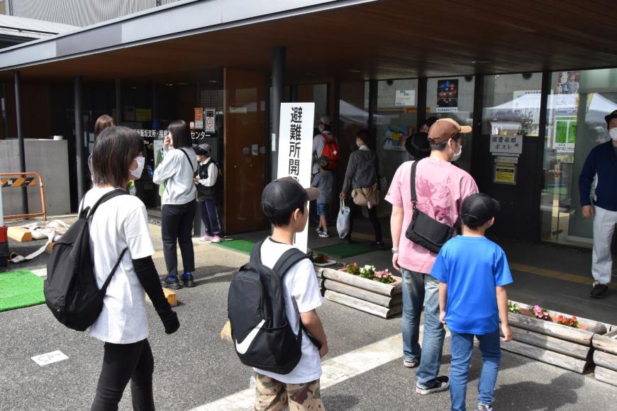 入口前に「避難所」と書かれた看板が立っている建物に避難をしようとしている住民の方達の訓練の様子の写真