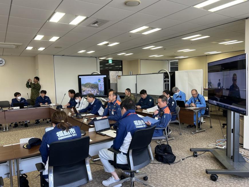 ホワイトボードやモニターが置かれた室内で災害対策本部設置訓練が行われている様子の写真