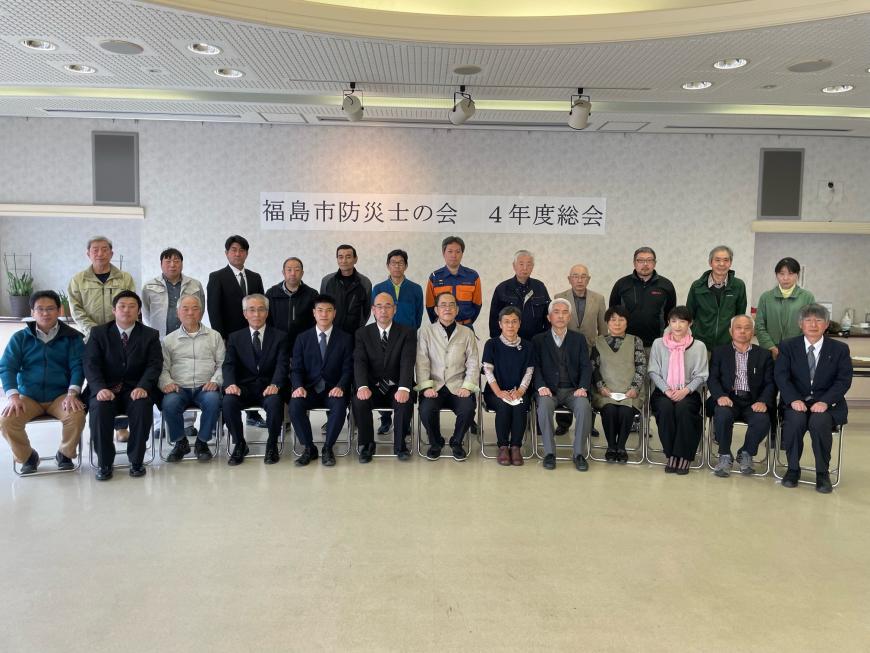 「福島市防災士の会 4年度総会」と壁に掲示され、参加者の皆さんが前列は椅子に座り、後に立って2列に並び写っている集合写真