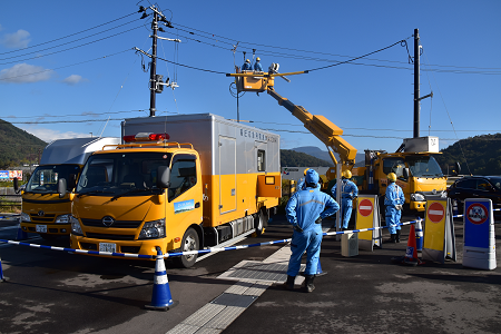 電線の通っている場所で、高所作業車に乗って作業をしており、道路に高圧応急用電源車と高所作業車など3台のトラックと青い作業福を着た方々が集まって作業を見守っている様子の写真