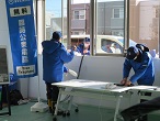 室内で作業を行う人が建物横に停めた車の中の男性とが窓越しにやり取りをしている様子の写真