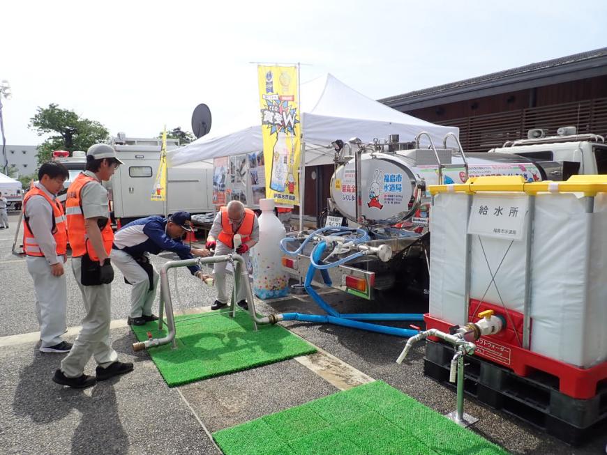 「給水所」と書かれた紙が貼られた四角いタンクが設置され、その横に設置された給水車につながった青いホースの先にある蛇口のような場所から水をコップに入れようとしている男性の写真