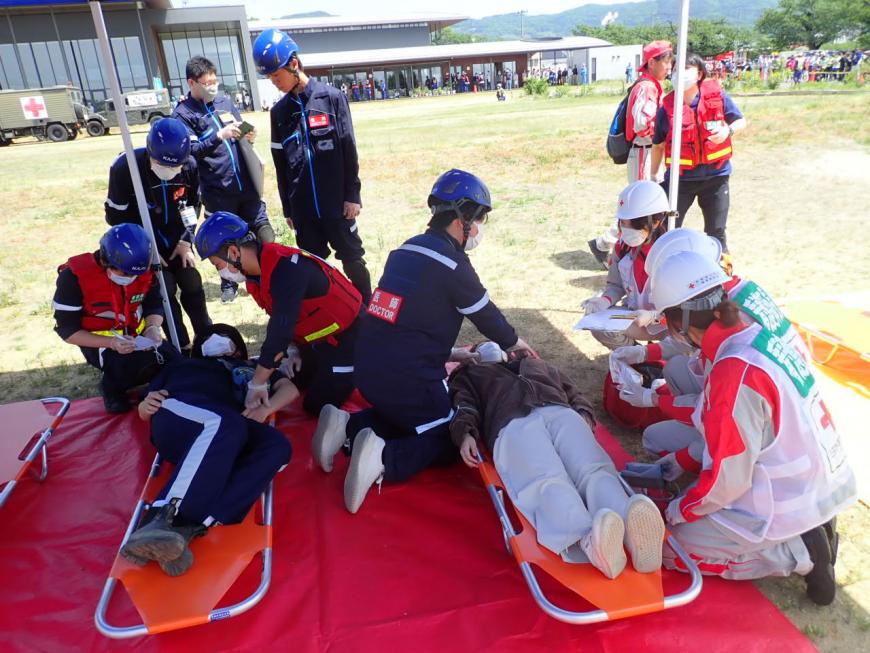地面に敷かれた赤いシートの上の担架に乗せられた2名の方の応急救護訓練を行っているDMATの方達の写真