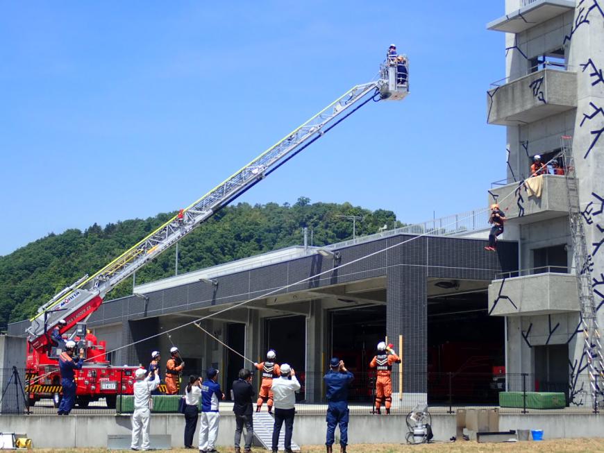 ひびが入った建物を模したコンクリート製の建物にロープが付けられ、ロープを使って建物から避難する訓練を行っている消防の方々の写真
