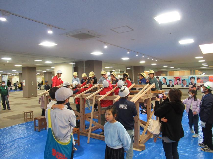 家の屋根の形に組まれた木材の反対側に黄色のヘルメット姿の子供たちが1列に並んでいる様子を周りの人たちが見ている写真