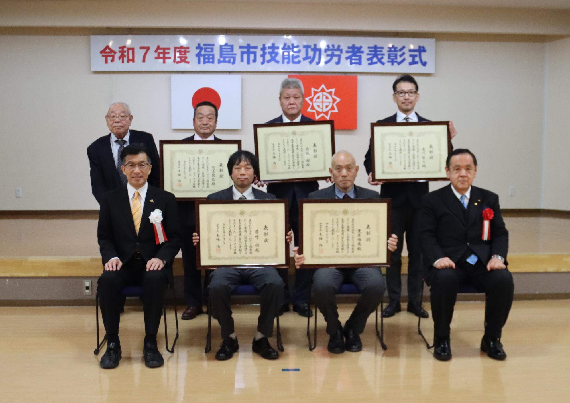 令和6年度 福島市技能功労者表彰式と書かれた横断幕と2枚の国旗が掲げられたステージで、胸元に胸章リボンを付けた8名の功労者と市長が並んでいる記念写真