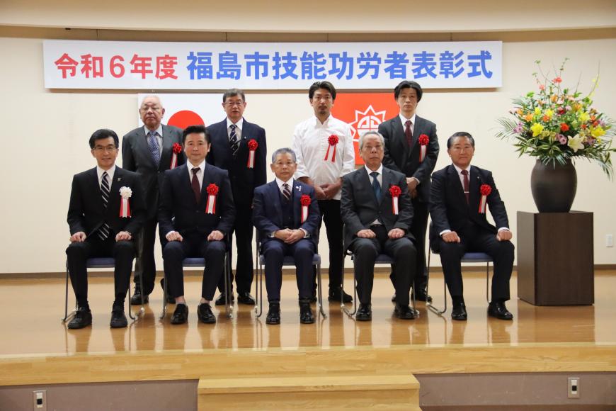 令和6年度 福島市技能功労者表彰式と書かれた横断幕と2枚の国旗が掲げられたステージで、胸元に胸章リボンを付けた8名の功労者と市長が並んでいる記念写真